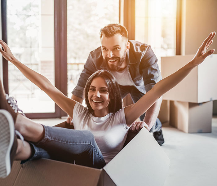 couple smilng together in their home pushing the woman while sitting in a box